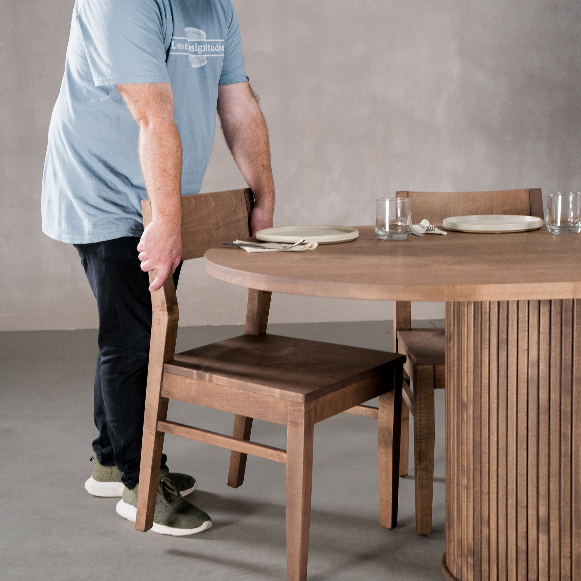 person pushing in a chair at a racetrack oval Maple table 42” wide x 84” long, finished in Coconut W on oval Como pedestal base